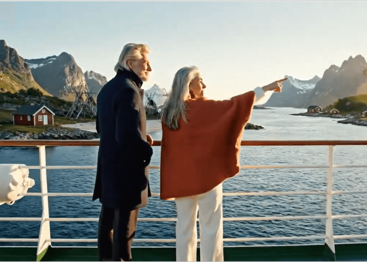 Guests on a cruise ship deck pointing at the Norwegian fjord landscape — red cabins, drying racks and snow-capped peaks